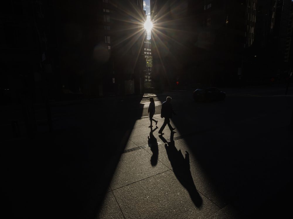 Two pedestrians walk through a beam of sunlight in downtown Toronto. The bright light of the Sun contrasts with the dark colours of buildings and the shadows of the pedestrians.