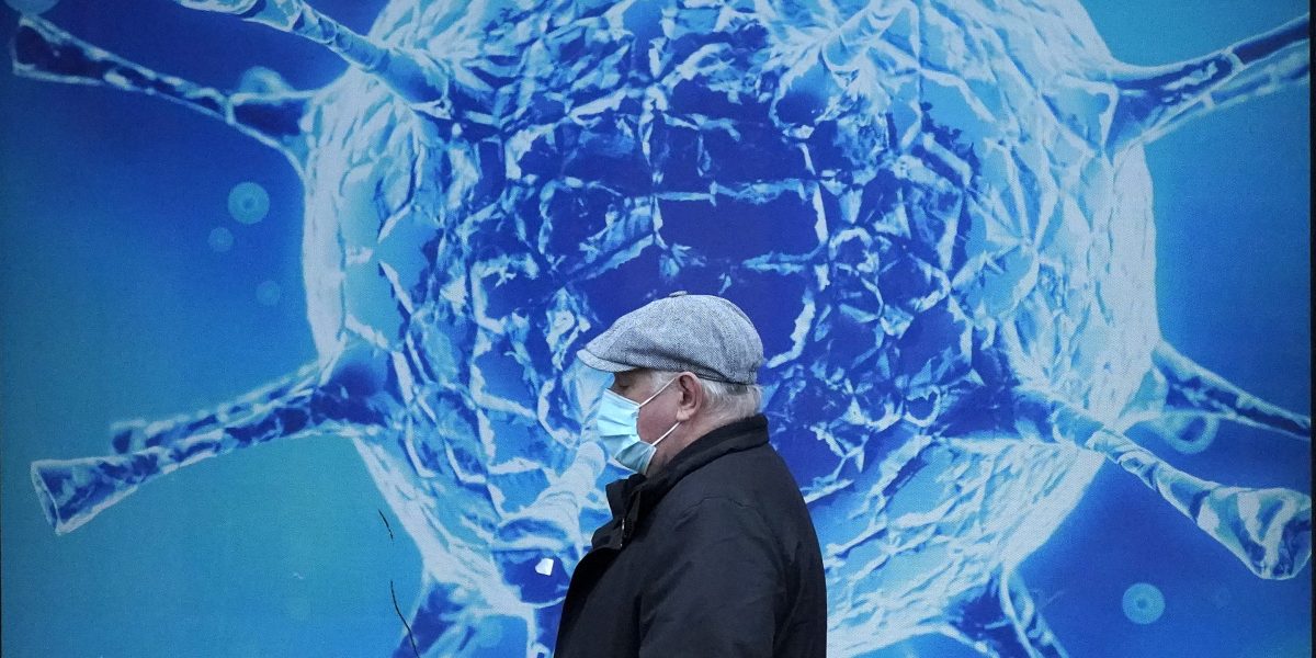 In the foreground, there is a person with a blue medical mask and a black jacket. There is a blue background, with an enlarged image of a SARS-CoV-2 virus particle.