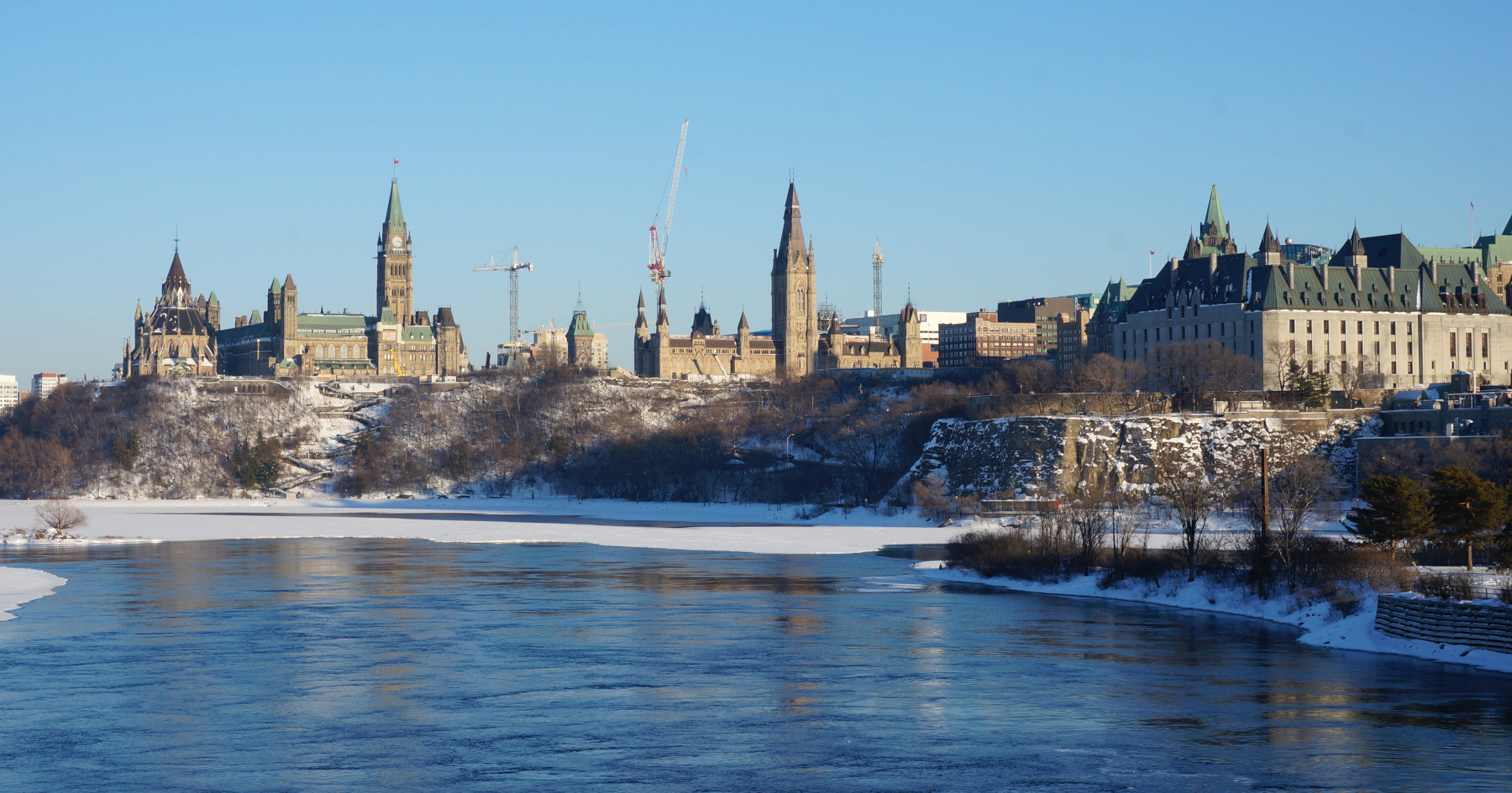 Photo of Parliament Hill, the Supreme Court, and the Ottawa River in Ottawa. There is snow on the ground and part of the river is covered with snow and ice. It’s a sunny day, with a blue sky.