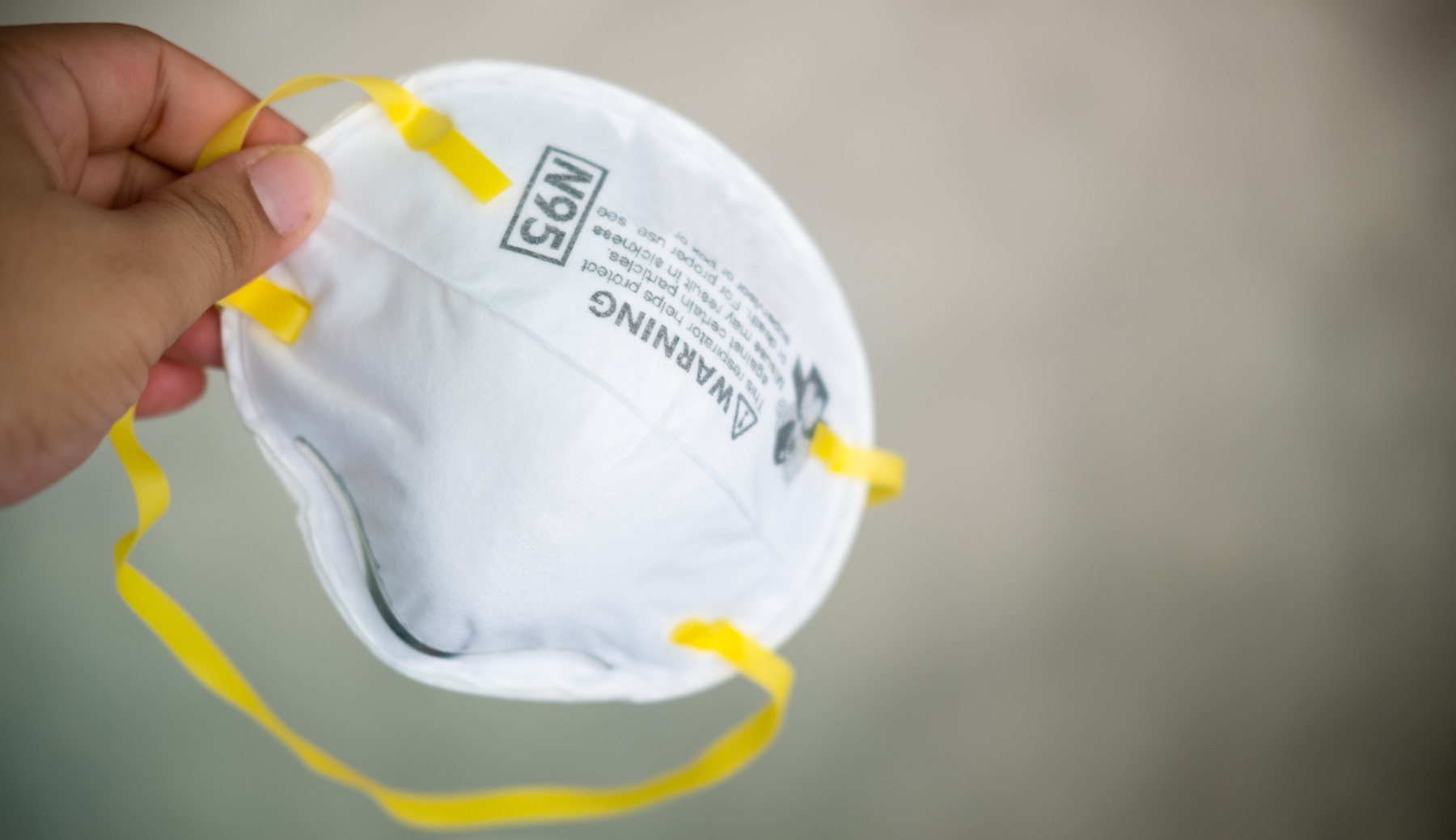 Hand holding a white N95 respirator with yellow headbands.