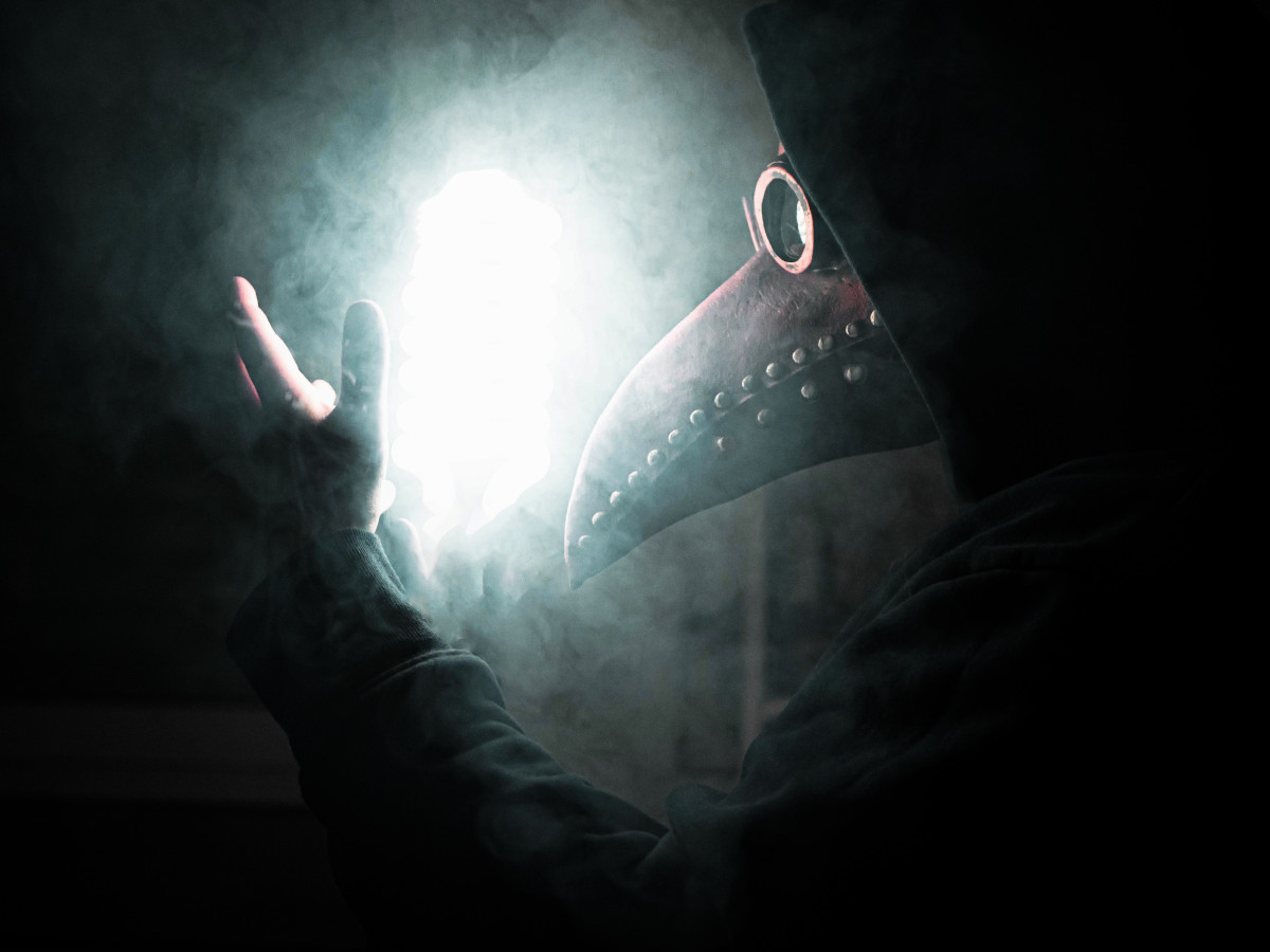 Image of a plague doctor wearing a plague mask and black clothes. The setting appears to be a smoky, dimly-lit room, with a bright light near the centre. Art by нυвιѕ тανєяη on Unsplash.