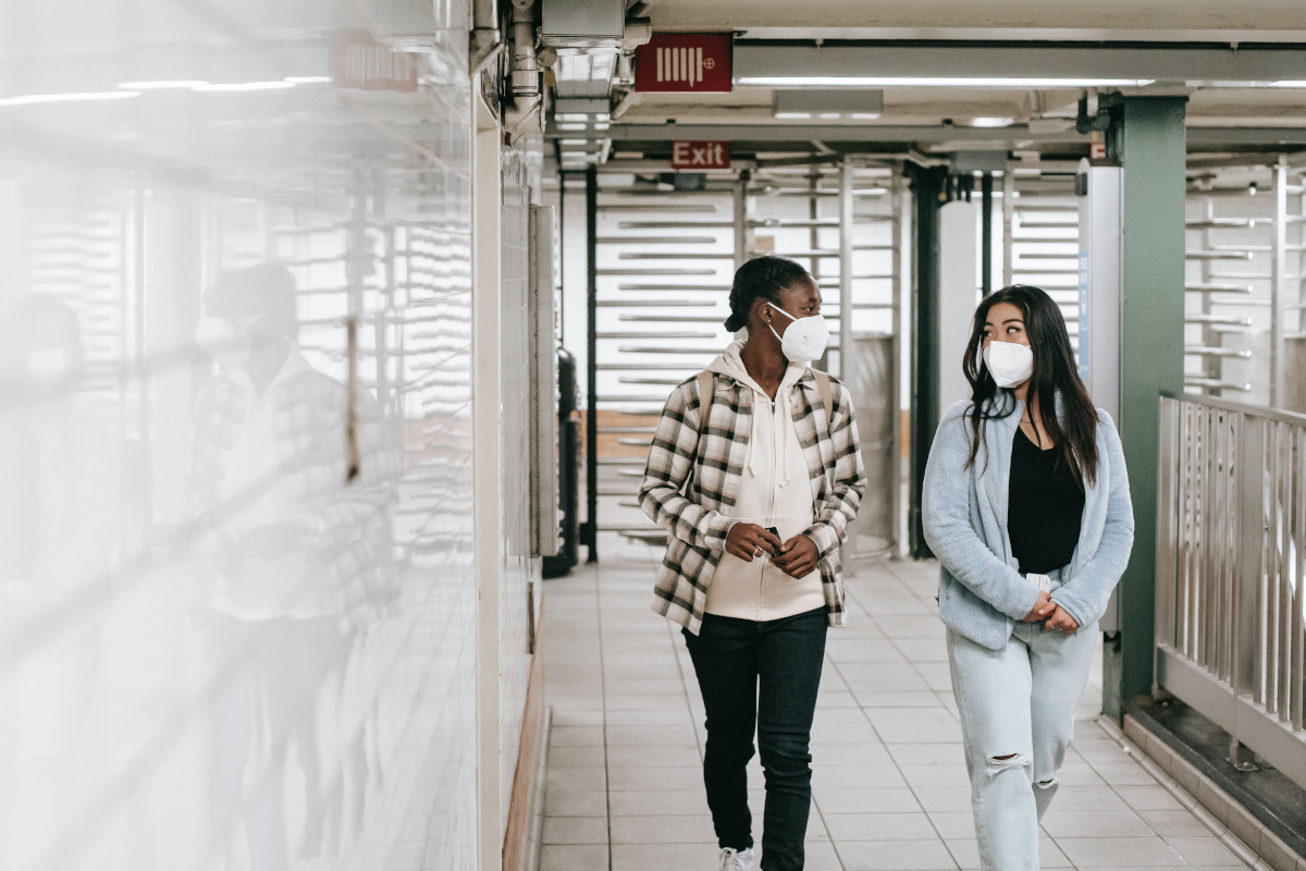Photo of two masked friends walking in a corridor in an underground public transit station. Photo by Charlotte May / Pexels.