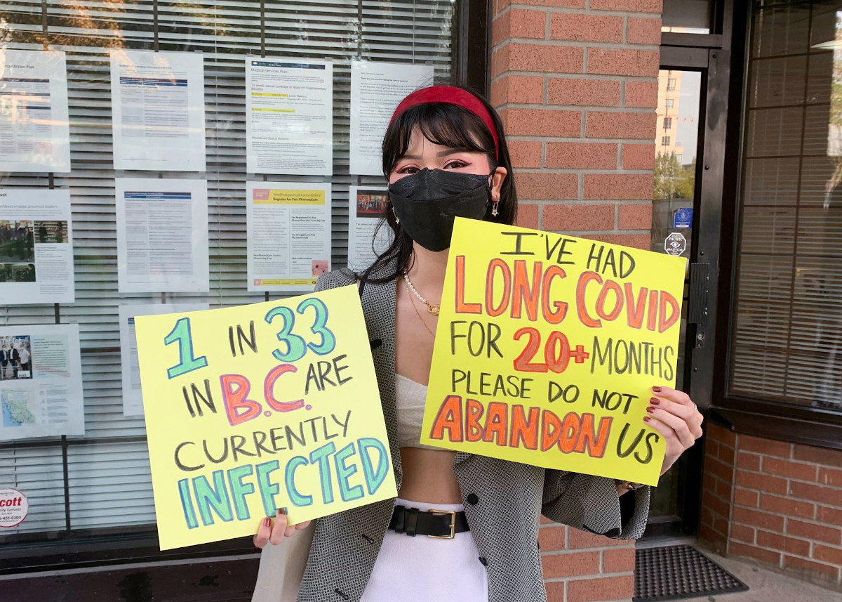 A young person with long black hair, a red headband and a black respirator holds two yellow signs reading: “1 in 33 in BC are currently infected” and “I’ve had long Covid for 20+ months. Please do not abandon us.” Photo: DoNoHarm BC.