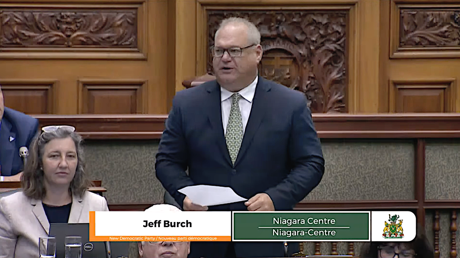 Screenshot of the broadcast of Question Period, on June 6, 2024. Jeff Burch, Member of Provincial Parliament (MPP) for Niagara Centre, is speaking. He has white and grey hair, glasses, a tie, and a navy blue suit. In the background, there are wood panels on the walls with intricate carvings.