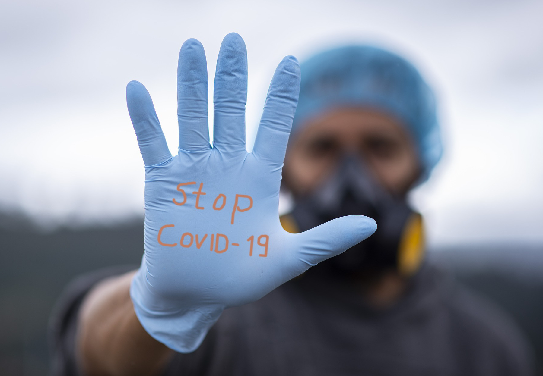 Photo of a person wearing an elastomeric respirator, with an outstretched, gloved hand reaching forward towards the viewer. On the light blue glove, the text reads: Stop COVID-19. With a bokeh effect, there is a sharp image of the glove, and the person’s face and respirator are blurred in the background. Photo: fernando zhiminaicela via Pixabay.